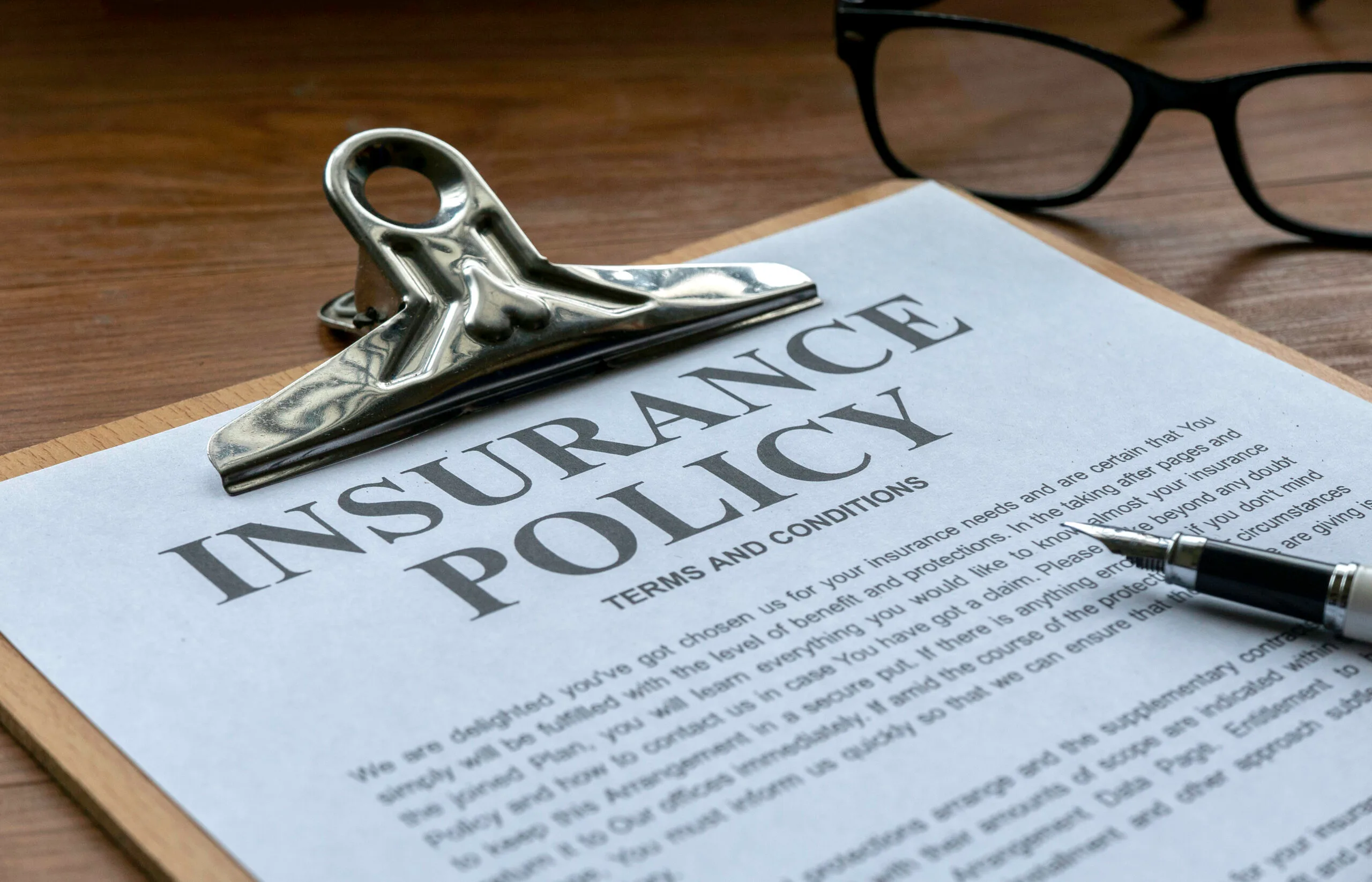 A clipboard holds an insurance policy document with the title in bold letters, next to a fountain pen and eyeglasses on a wooden desk.