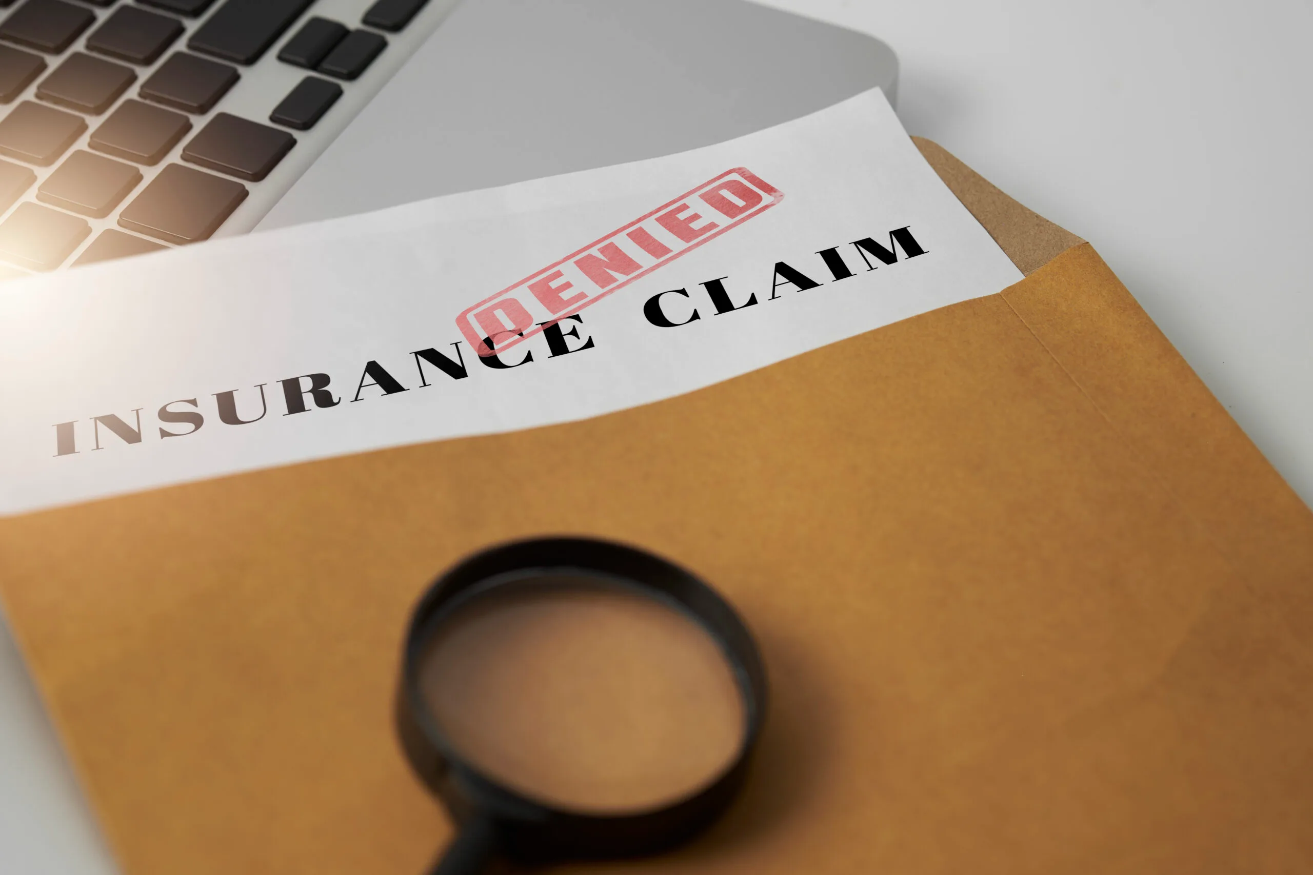 A document labeled “Insurance Claim” with a red “Denied” stamp lies on a folder beside a laptop and magnifying glass.