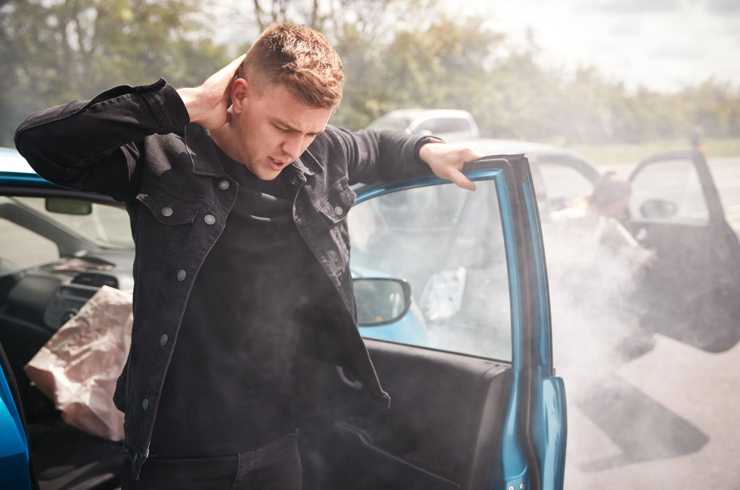 Injured man holding his neck after a car accident, standing next to a smoking vehicle with airbags deployed.