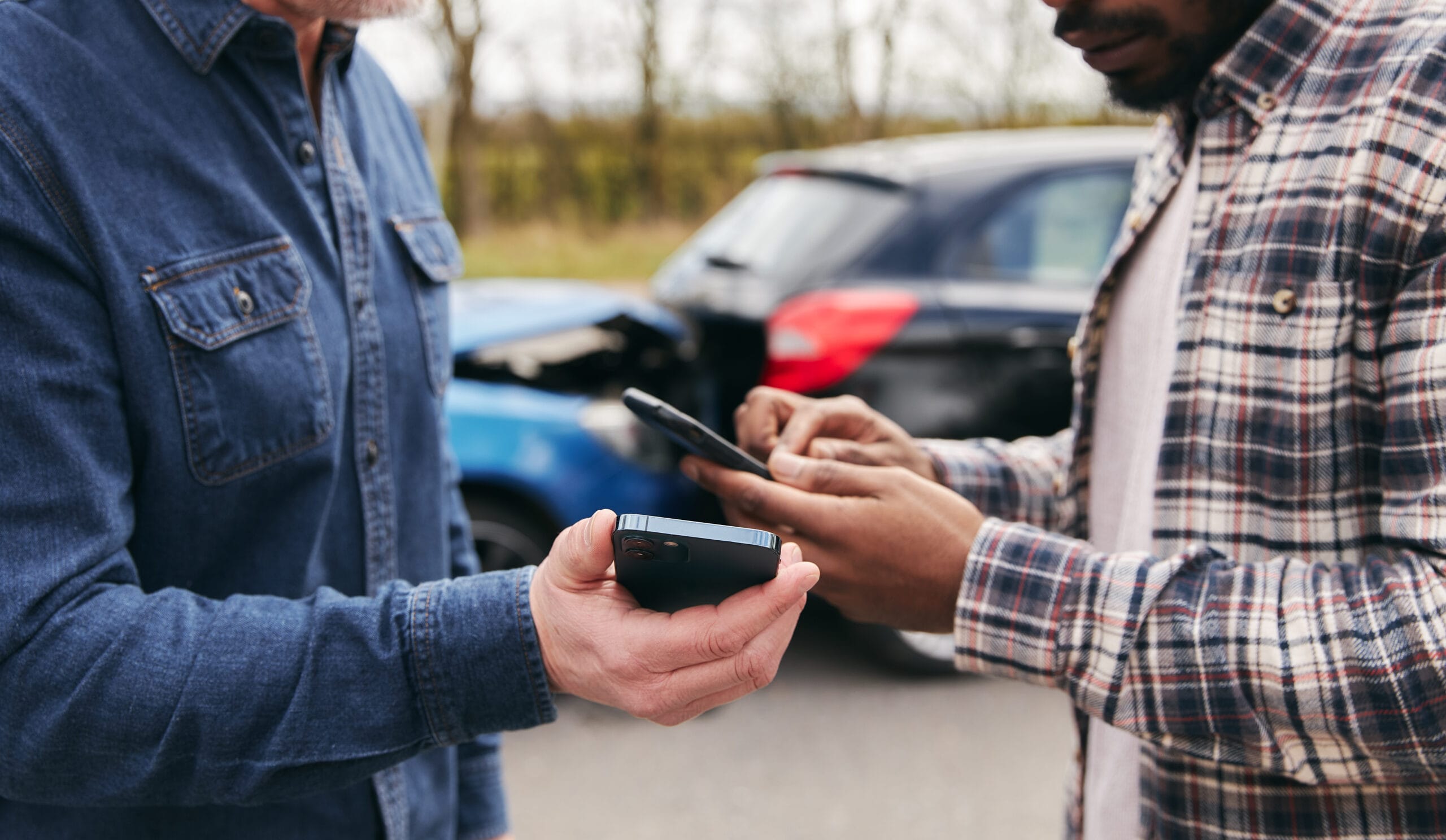 Two drivers exchange information on their phones after a car accident, with damaged vehicles visible in the background on a roadside.