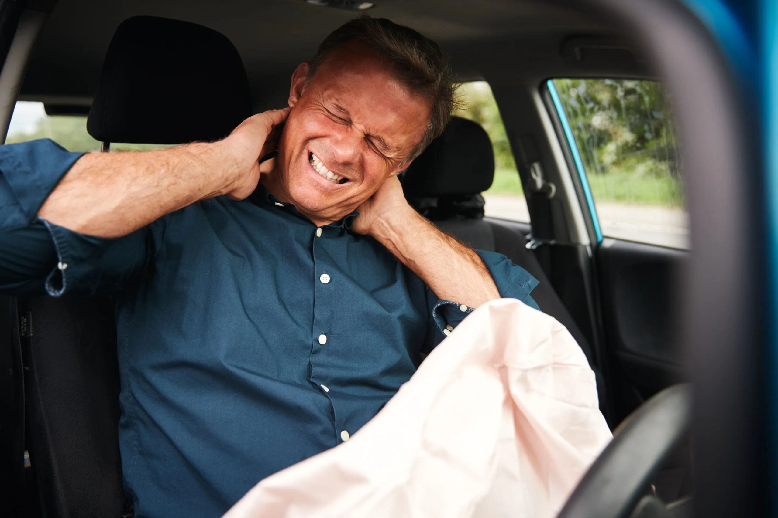 A man sitting in the driver’s seat of a car holds his neck in pain after an accident, with the airbag deployed in front of him.