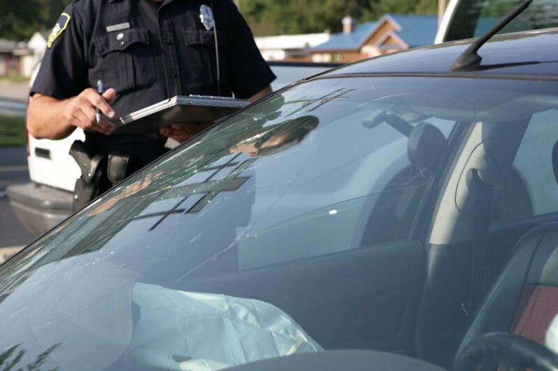 A police officer writes notes on a clipboard beside a car with a cracked windshield and deployed airbag after an accident.