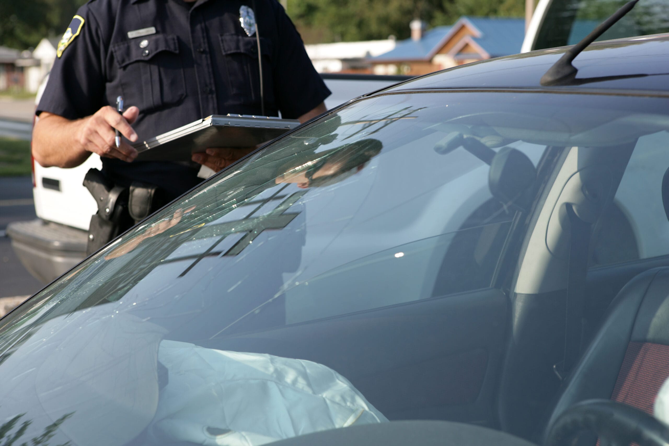 A police officer writes notes on a clipboard beside a car with a cracked windshield and deployed airbag after an accident.