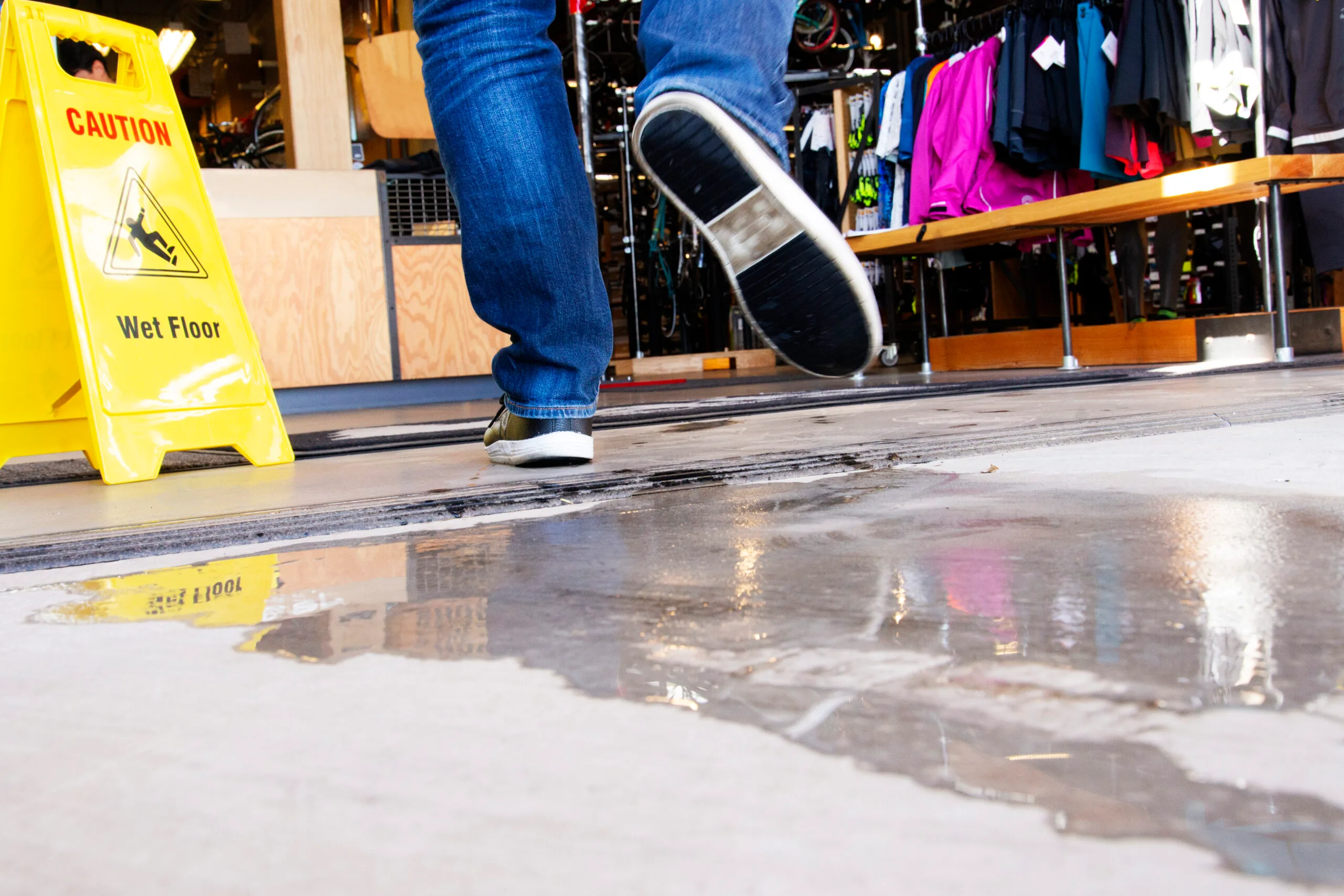 A person walking over a puddle on a store floor near a yellow “Caution: Wet Floor” sign.