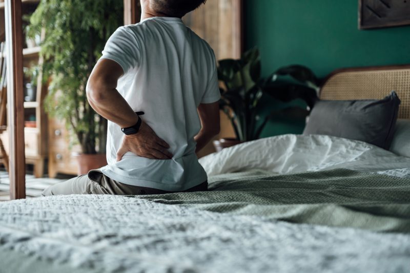 Injured man sitting on the edge of a bed holding his back.