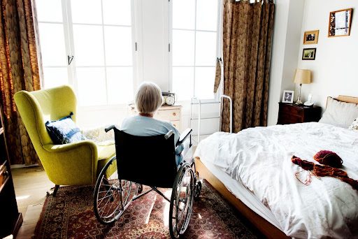 Elderly woman in a wheelchair facing the windows in her nursing home room.