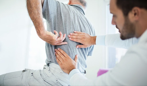 An elderly man indicates a pain point on his back while a doctor examines the area.