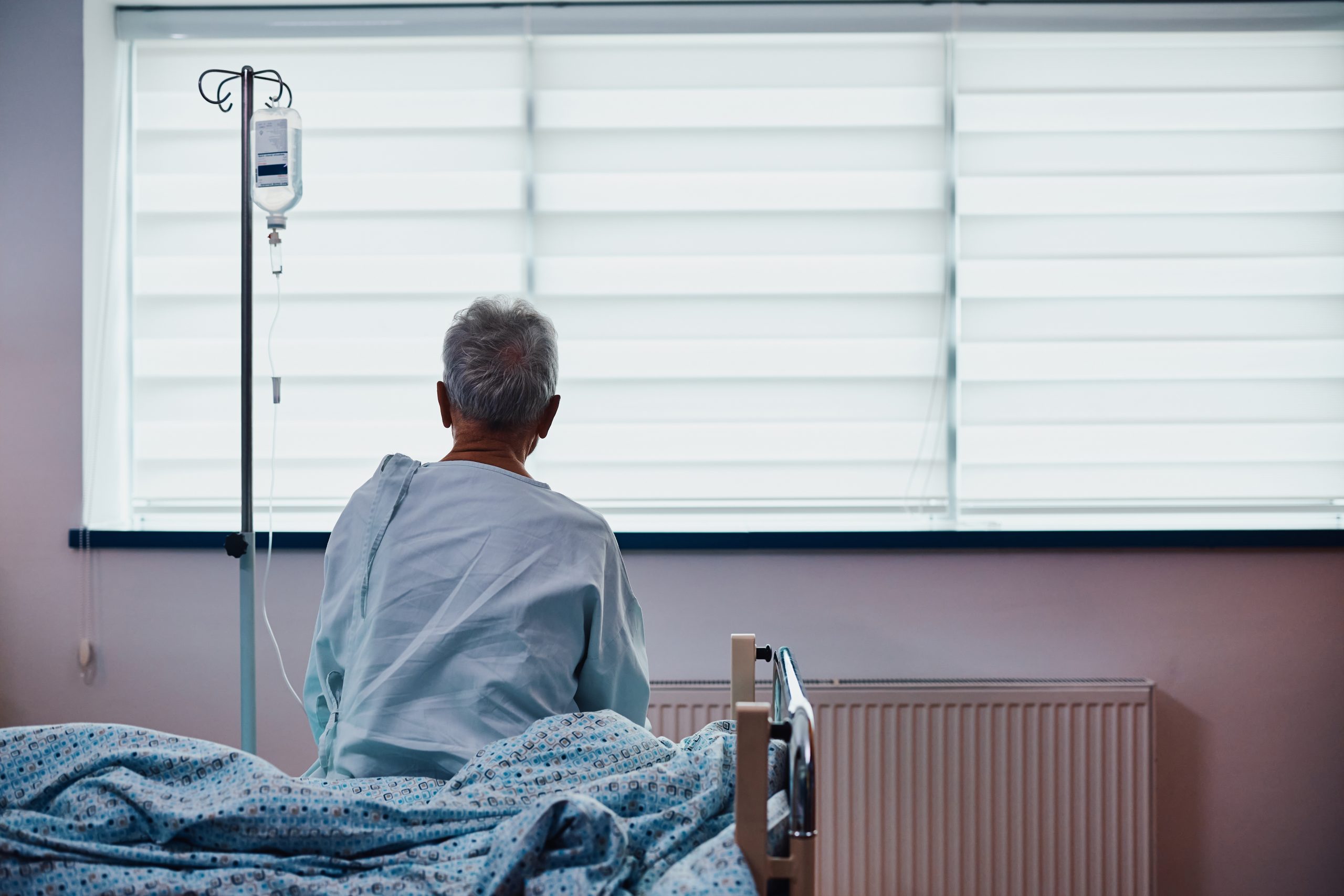 An elderly person in a hospital gown sits on the edge of a bed, facing a window with an IV drip stand.