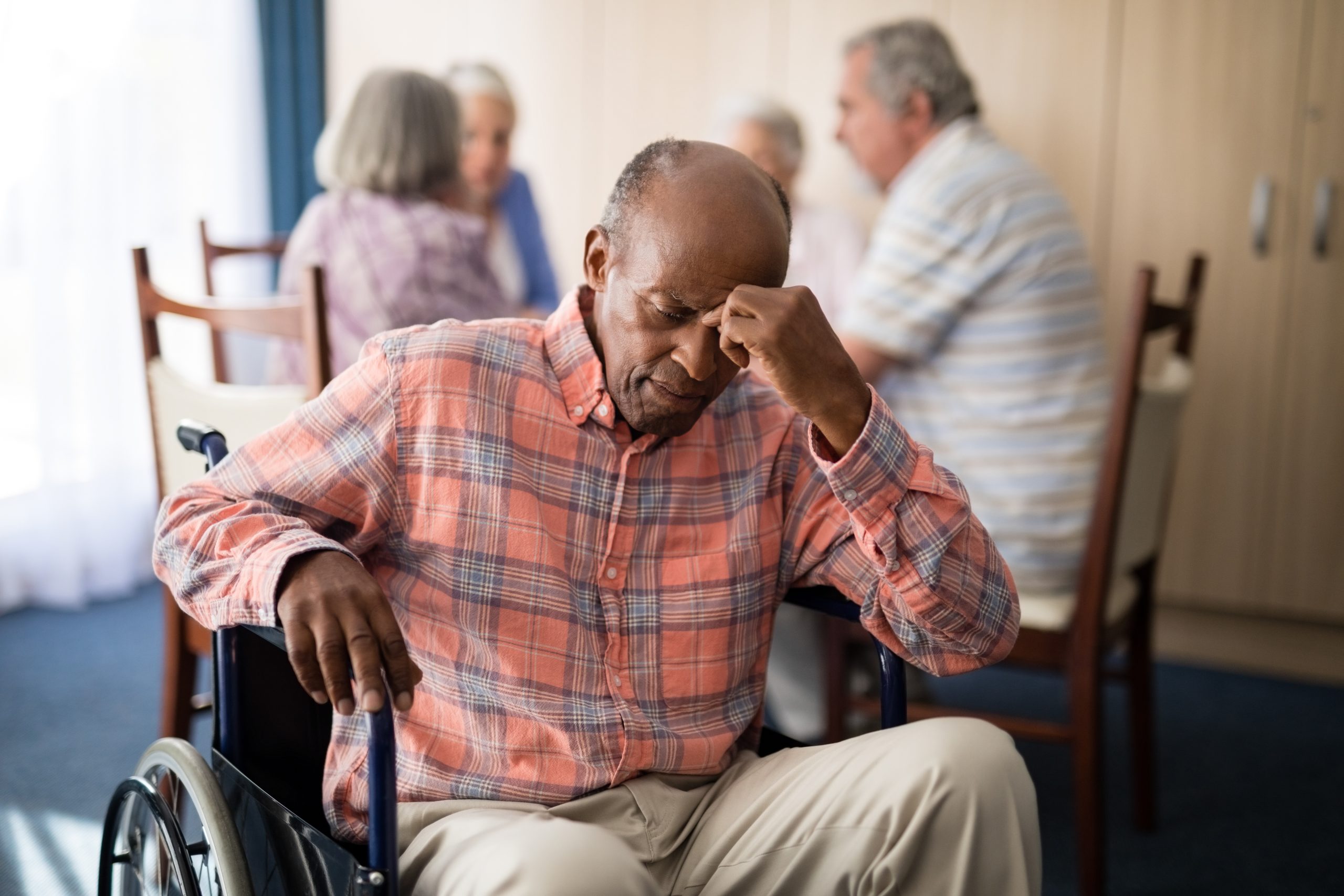 An elderly man in a wheelchair sitting with his head resting on his hand with nursing home residents seated at a table in the background.