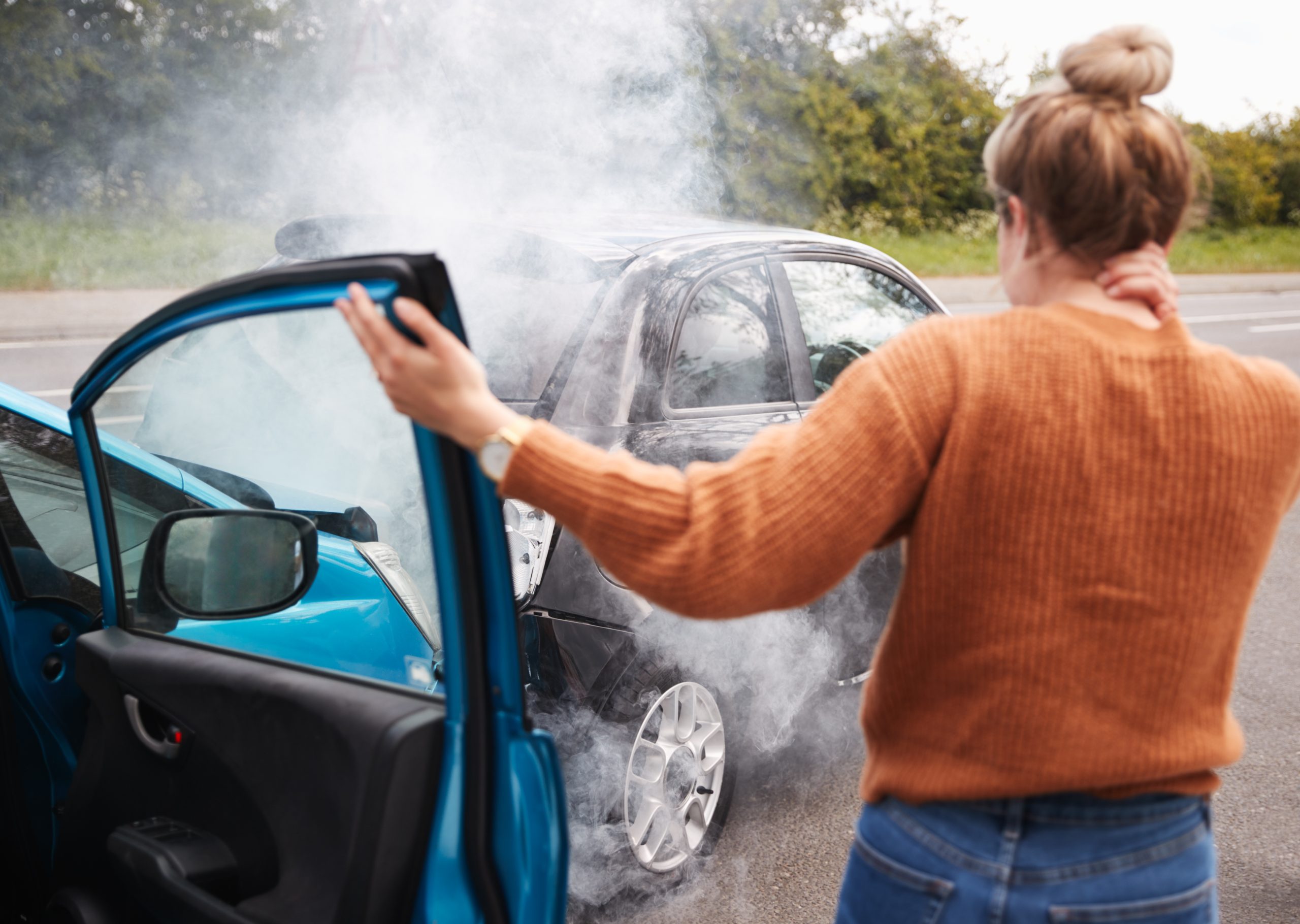 A woman stands outside her vehicle holding her neck and examining the damage to her blue car after a collision with a black car. Smoke is coming from the vehicles.