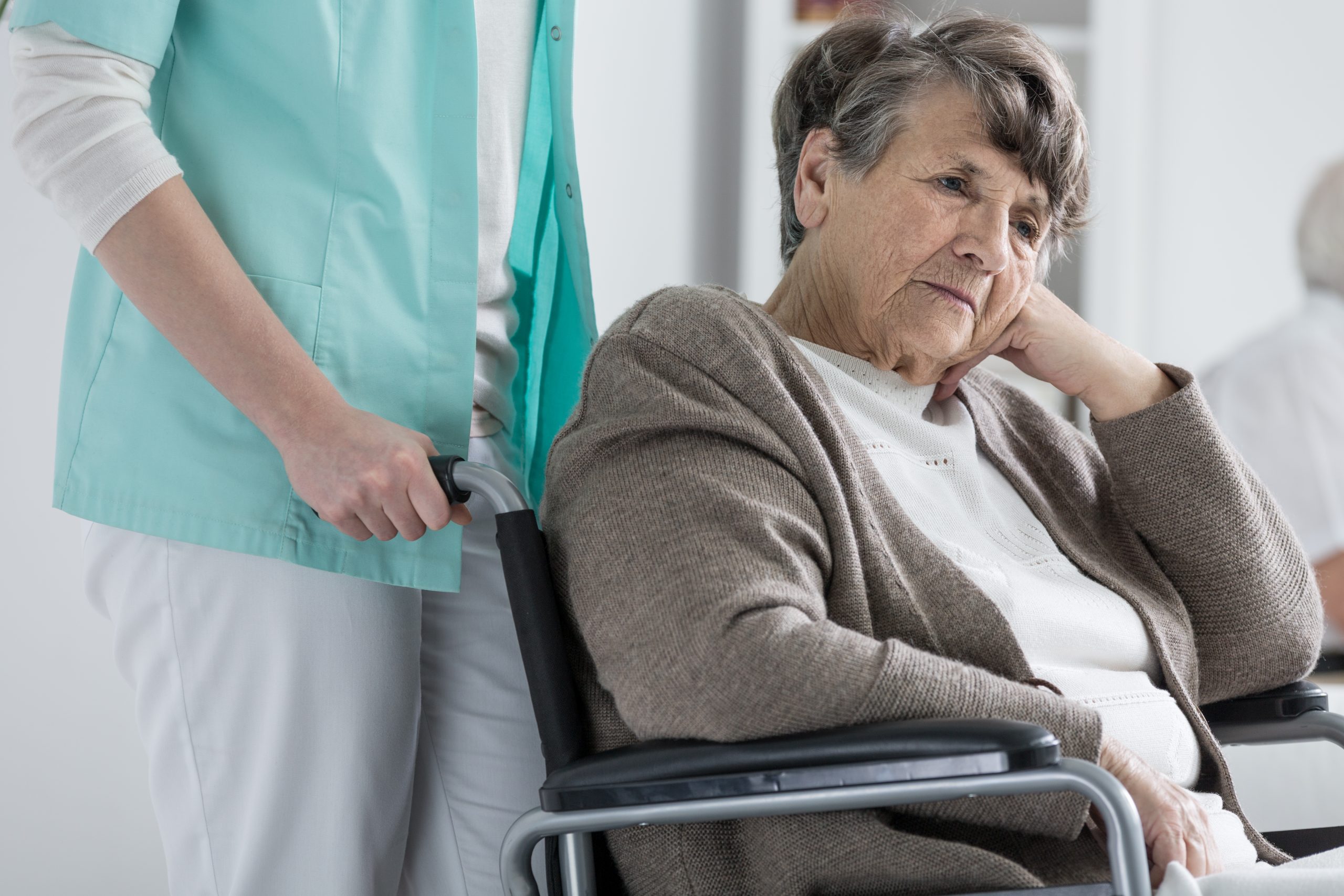 An elderly woman sits in a wheelchair, looking sad and contemplative, while a caregiver stands beside her, holding the wheelchair handles.