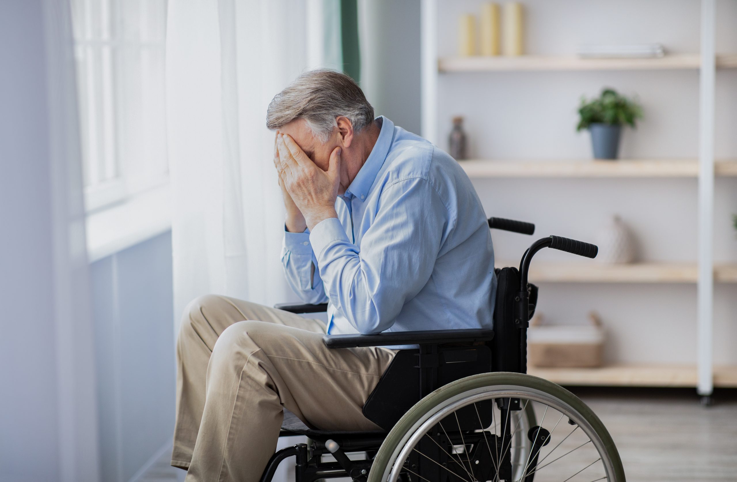 An elderly man in a wheelchair covers his face with his hands, appearing distressed.