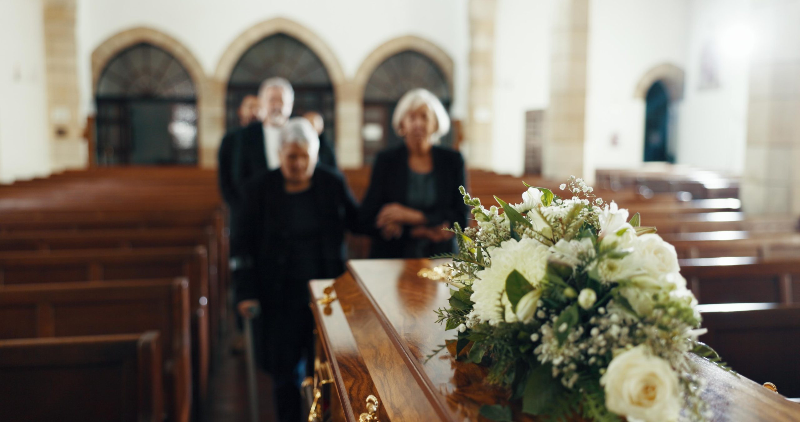 A group of mourners in a church walking towards a coffin adorned with white flowers.