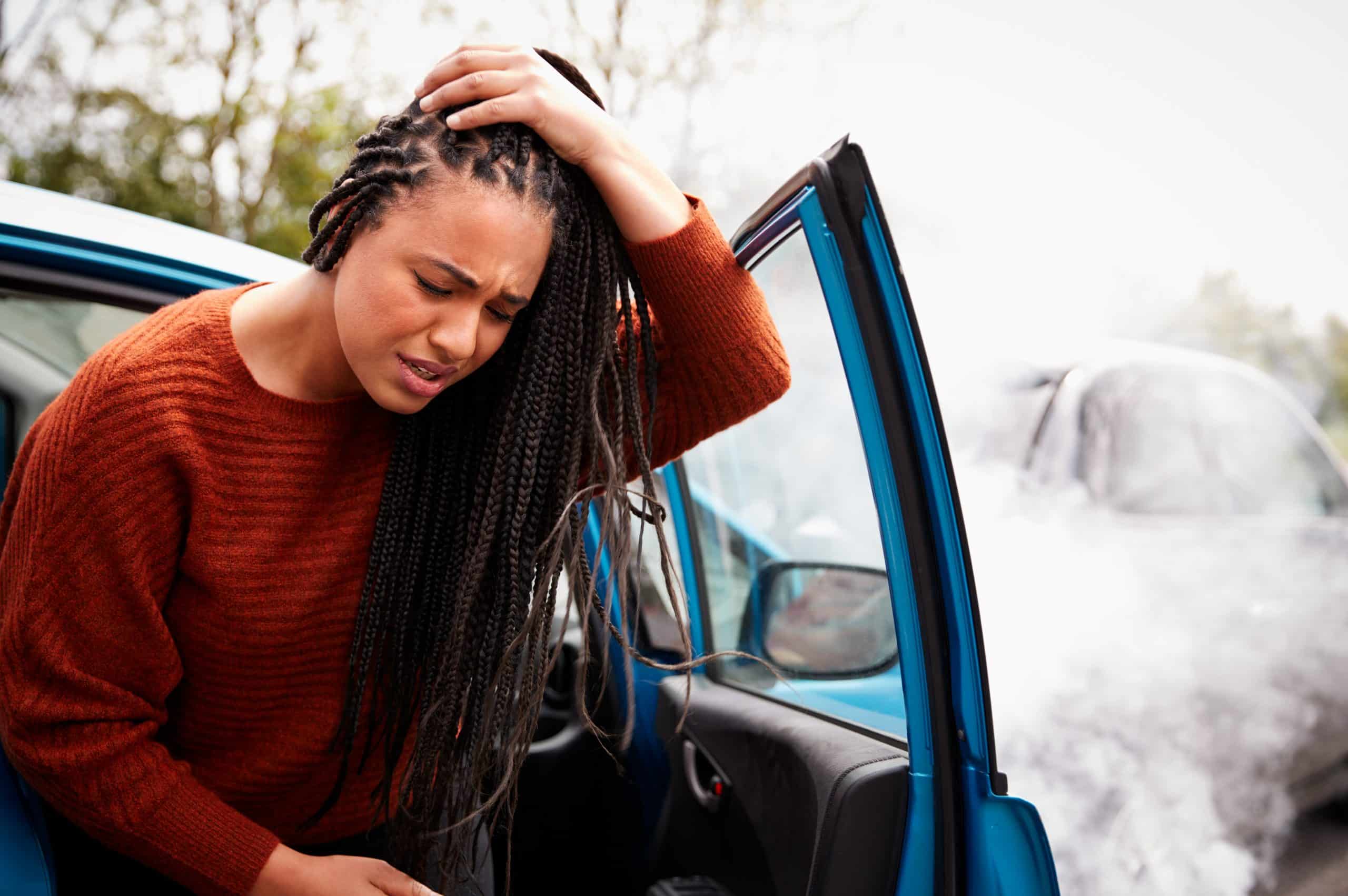 A woman holding her head in pain as she steps out of a damaged car after an accident. Another car in the background is obscured by smoke.