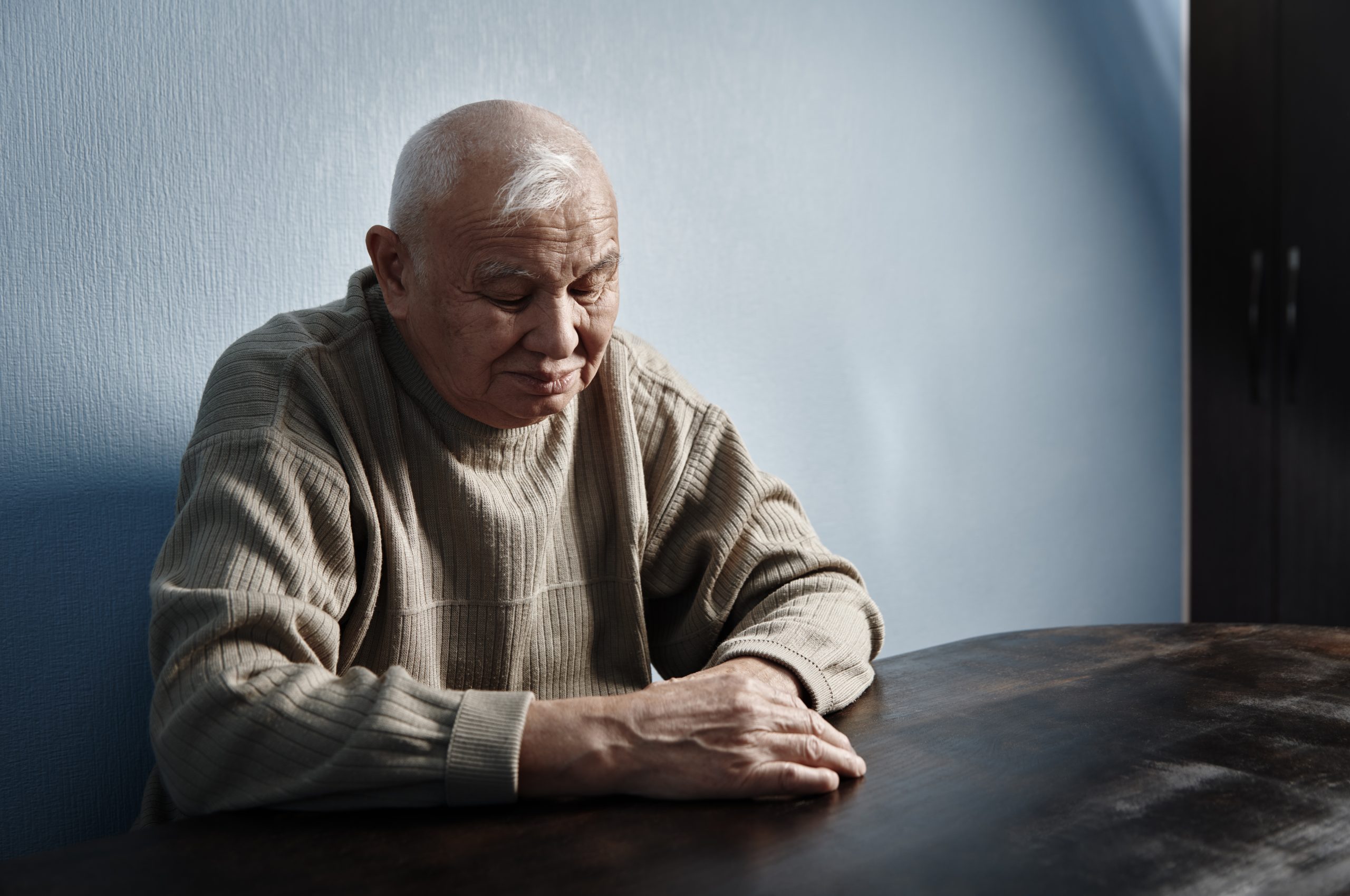 A senior man sitting alone at a table, looking down at his hands resting in front of him.