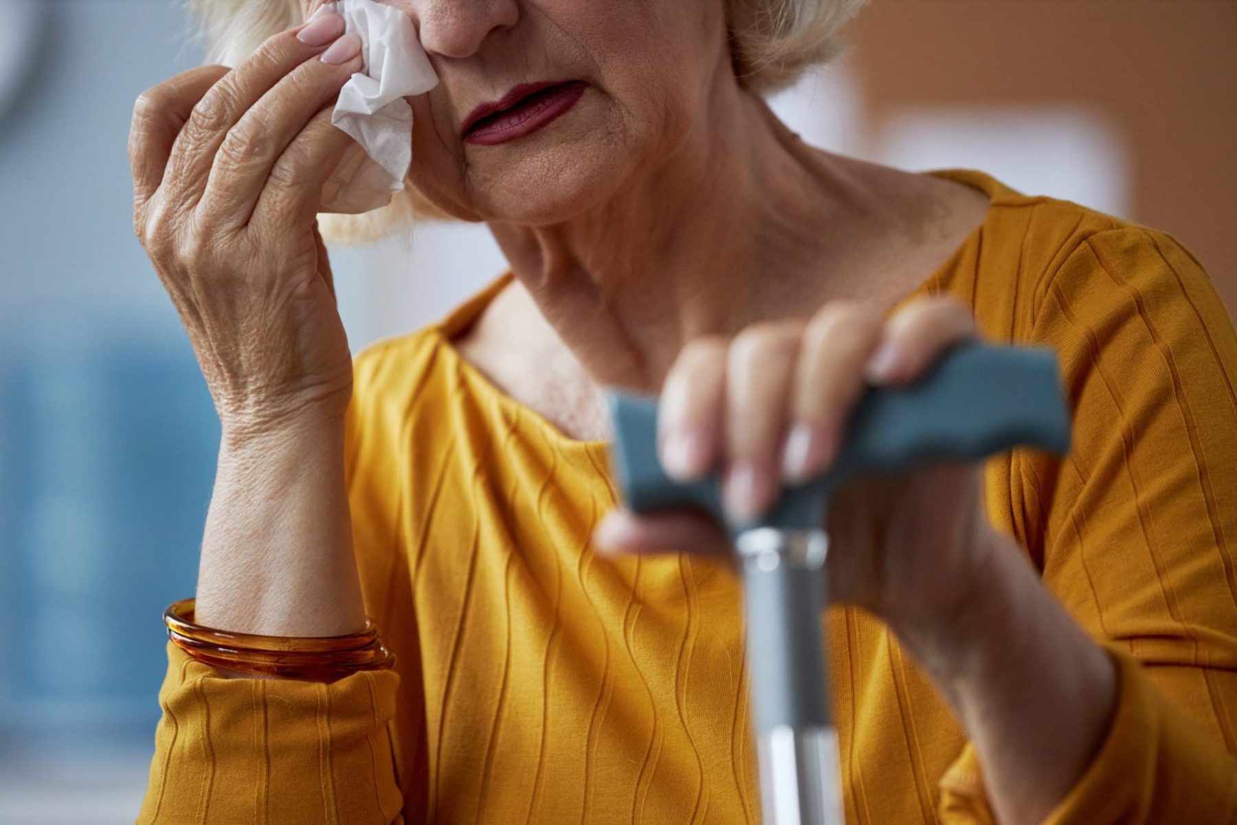 Close-up of an elderly woman wiping a tear with a tissue and holding a cane.