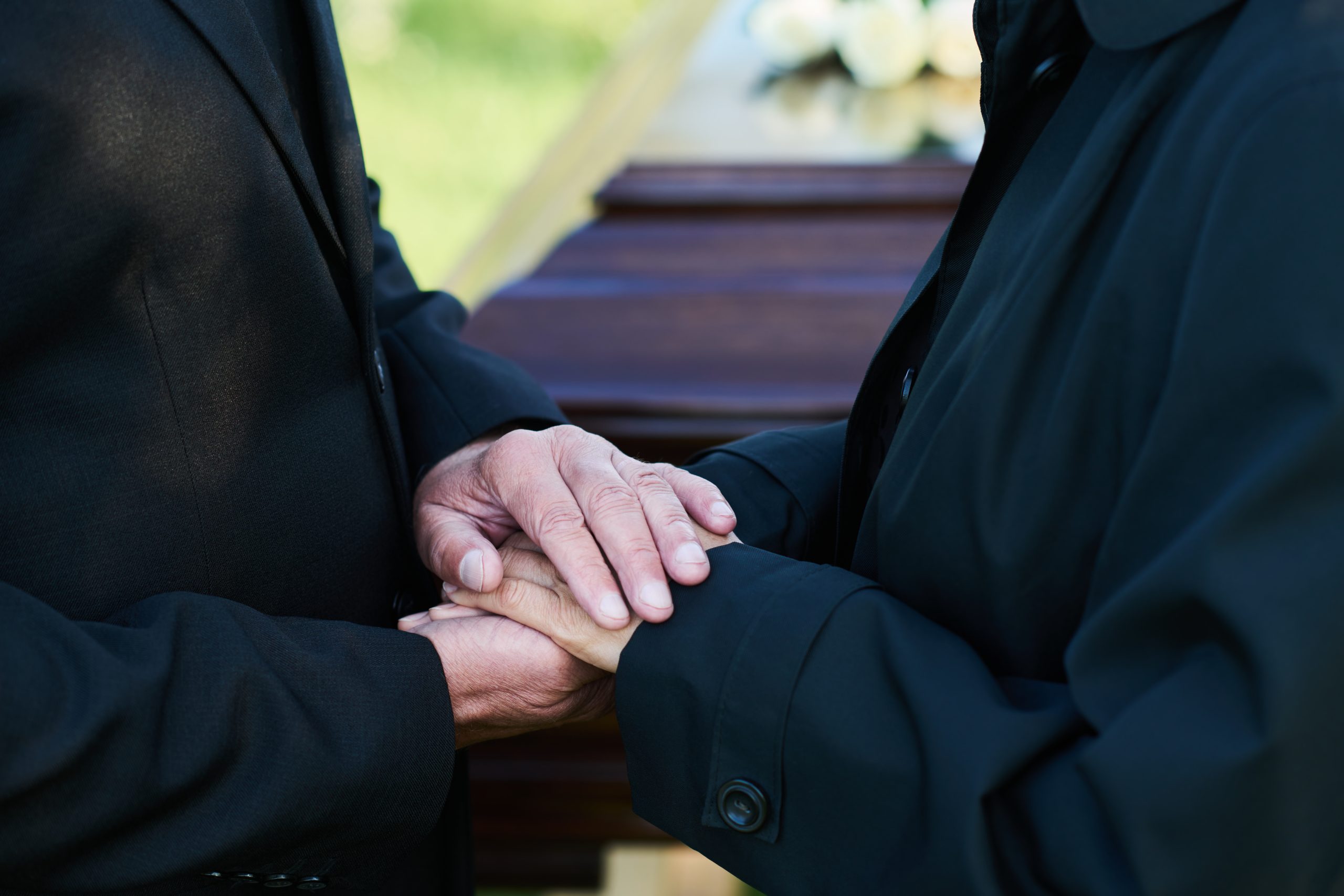 Close-up of two people in black clothing holding hands in front of a casket during a funeral.