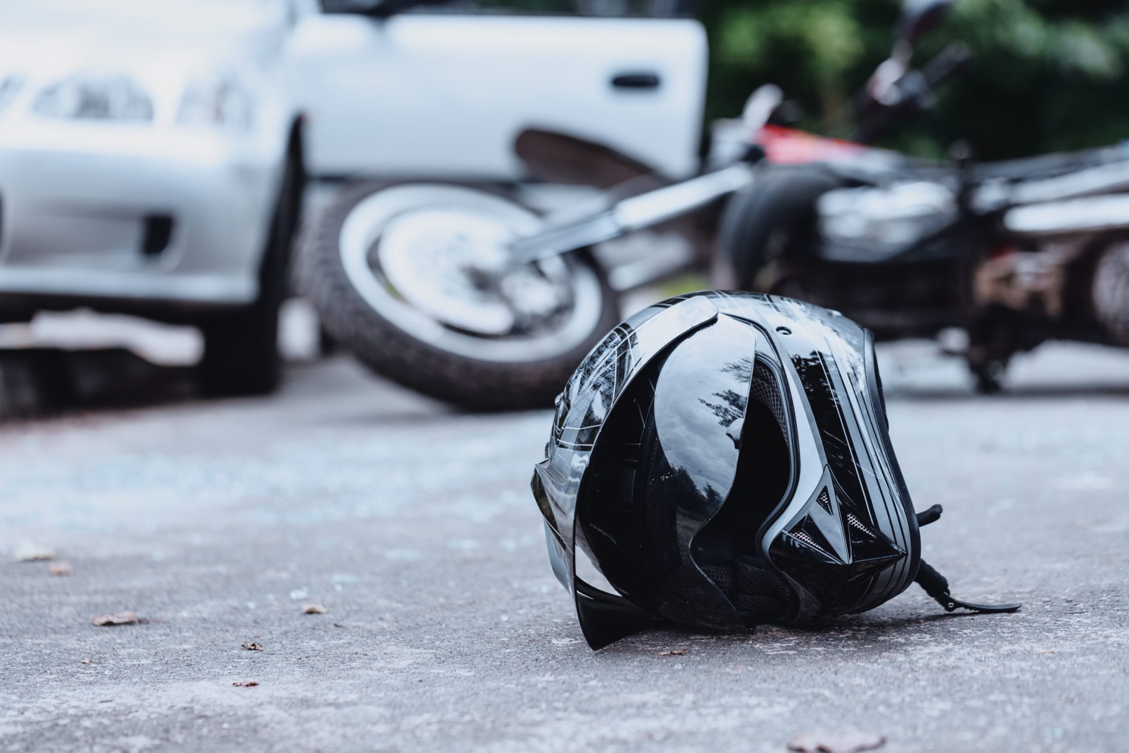 Close-up of a motorcycle helmet on the ground with a damaged motorcycle and car in the background.