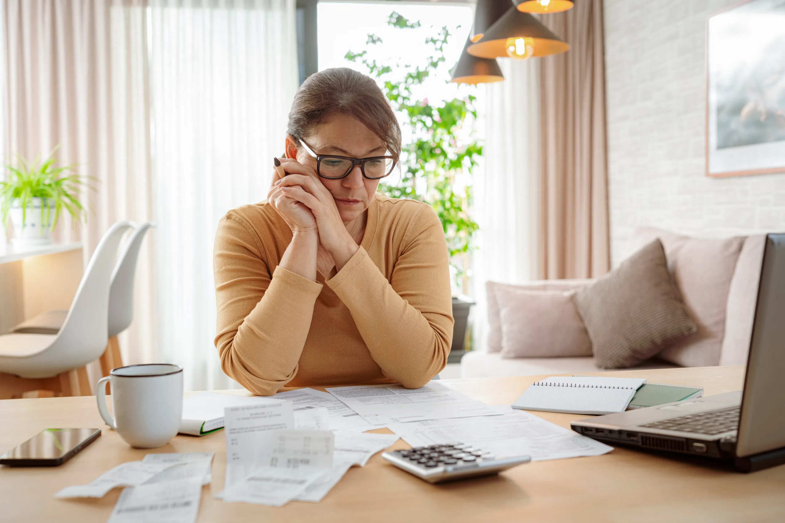 Concerned woman reviewing bills and financial documents at home with a calculator and laptop.