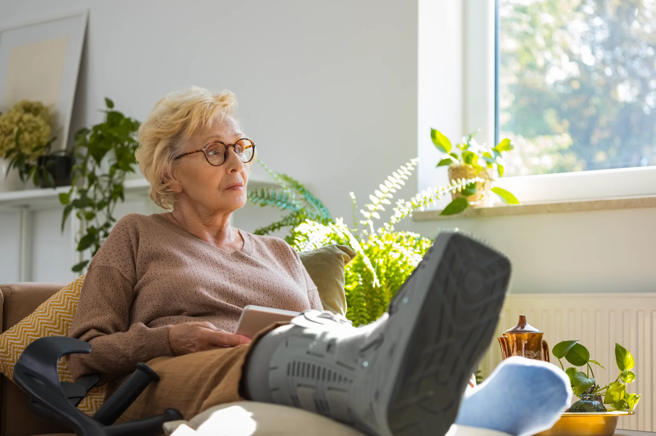 An elderly woman sits on a couch with her leg elevated in a medical walking boot.